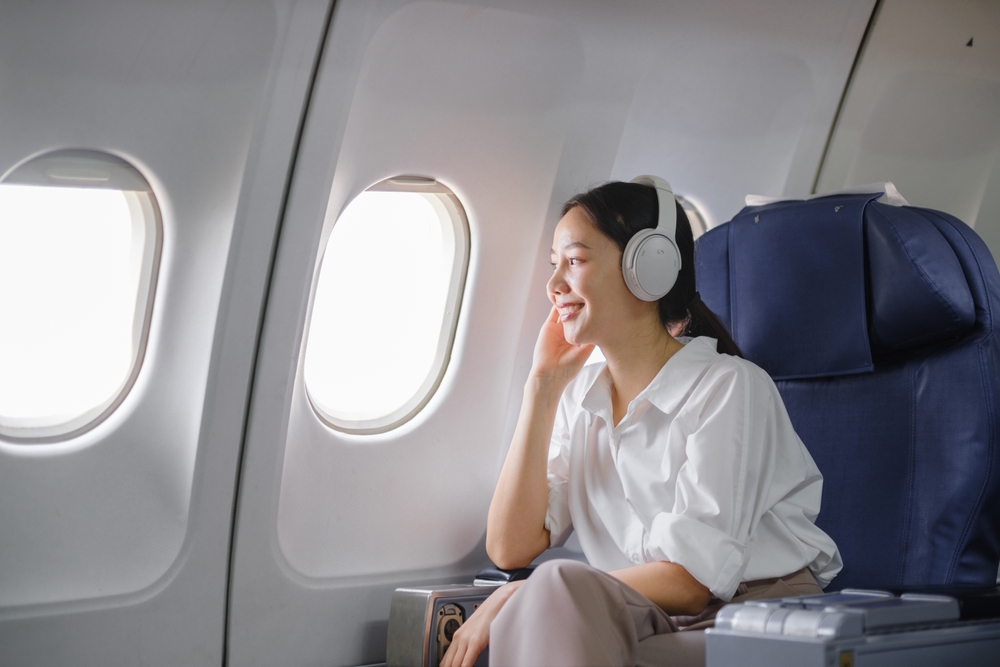 A woman wearing headphones smiles while looking out the window on an airplane. She is seated in a comfortable seat, dressed in a white shirt, and appears relaxed during her flight.