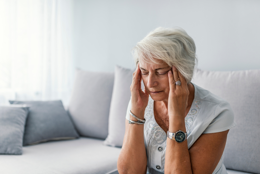 An older woman with short gray hair sits on a couch, eyes closed, and presses her fingers to her temples, appearing to have a headache or be in pain.