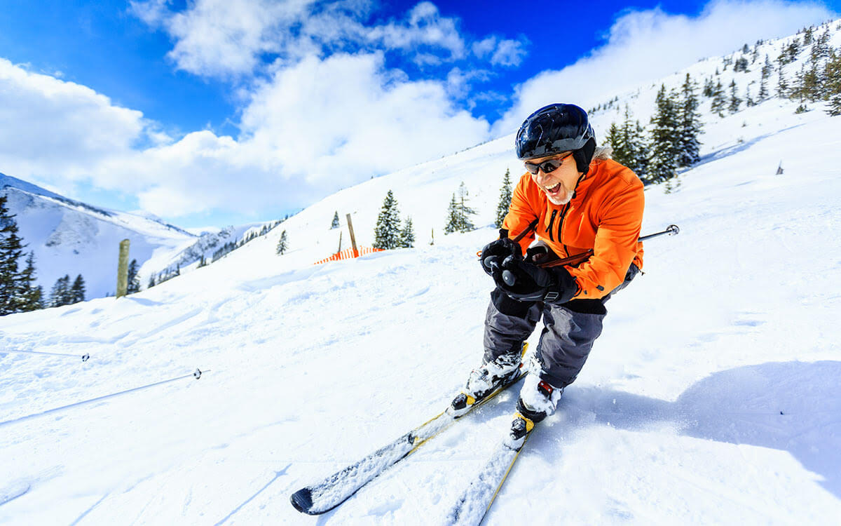 A person in an orange jacket and black helmet is skiing downhill on a snowy mountain, smiling and holding their ski poles, with pine trees and a bright blue sky in the background.